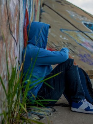 Photo of young person in blue hoodie sitting against wall with graffiti